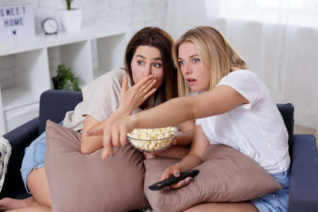 two terrified female friends watching news or scary movie on tv at homeの写真素材