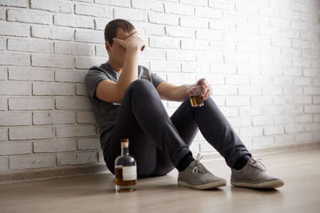 young man suffering from alcoholism, guy sitting on the floor with glass and whiskey bottle over white brick wallの写真素材