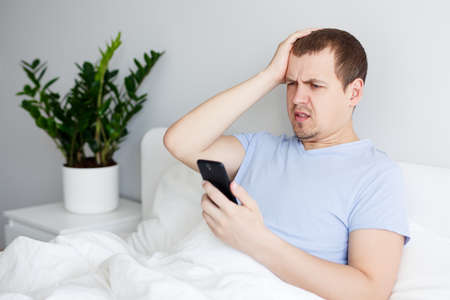 portrait of young upset man using smartphone in bed