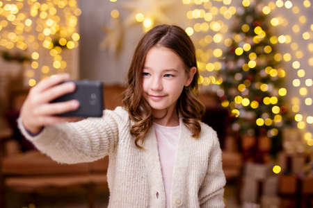 Christmas and new year concept - cute little girl taking selfie photo with smartphone in decorated living room with Christmas tree and lightsの写真素材