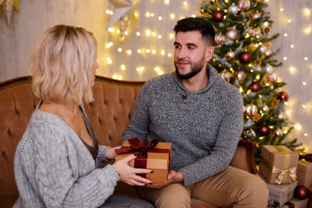 Christmas and love concept - handsome man giving Christmas present to his girlfriend in living room with decorated Christmas tree and lightsの写真素材