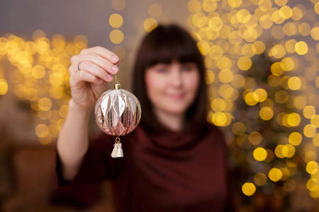 Christmas concept - portrait of beautiful woman decorating Christmas tree in decorated living room with holiday garland lights, focus on ballの写真素材