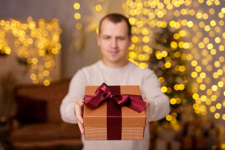 young handsome man giving gift box over christmas background with Christmas tree and led lights, focus on presentの写真素材