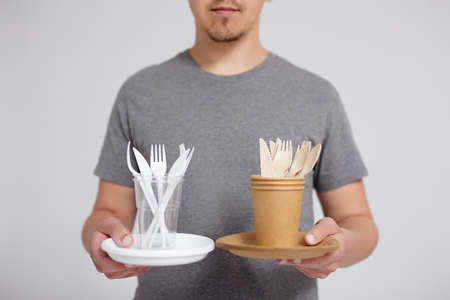 zero waste and eco friendly concept - young man comparing plastic with wooden and paper tableware over grey backgroundの写真素材