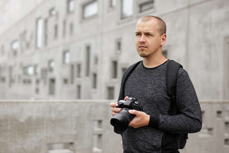 portrait of thoughtful male photographer posing with modern dslr camera over concrete building...