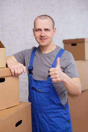 moving day or postal delivery concept - loader or deliveryman thumbing up over white brick wall with...