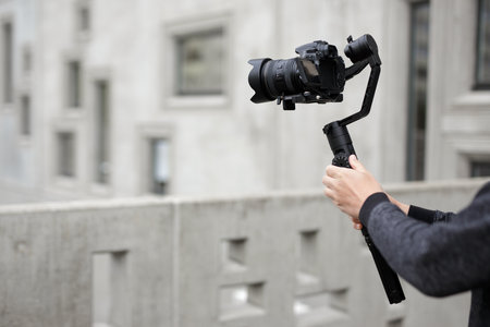 filmmaking, hobby and creativity concept - modern dslr camera on 3-axis gimbal in male hands over concrete building backgroundの写真素材