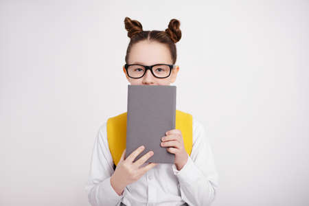 portrait of cute girl in school uniform and eyeglasses covering face with book over white background with copy spaceの写真素材
