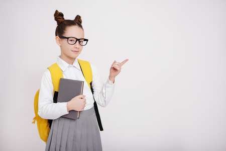 cute pretty girl in school uniform and eyeglasses with backpack and book pointing at something over white backgroundの写真素材