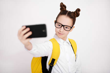 portrait of cute girl in school uniform and eyeglasses taking selfie photo with smart phone over...