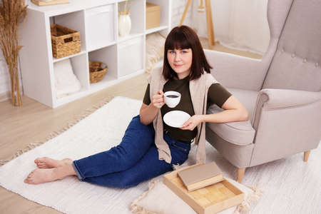 cute young woman sitting on carpet and drinking coffee in bright living room