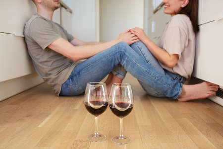 close up of glasses of red wine and young couple in love sitting on the floor in the kitchenの写真素材