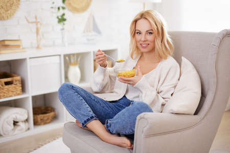 breakfast concept - happy young beautiful woman eating corn flakes in living room