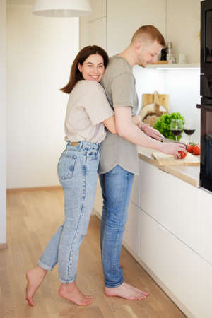 full length portrait of young woman embracing her boyfriend cooking dinner in modern kitchenの写真素材