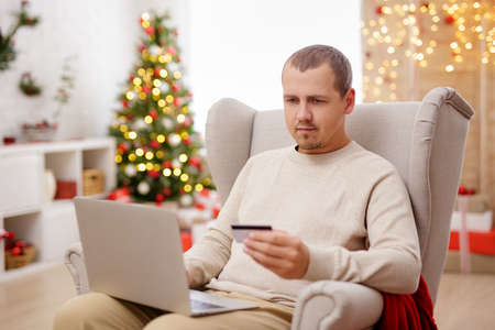 Young man with laptop doing online shopping in cozy christmas interiorの写真素材