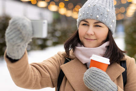 Close up of woman in winter clothes with coffee doing selfie in christmas marketの写真素材