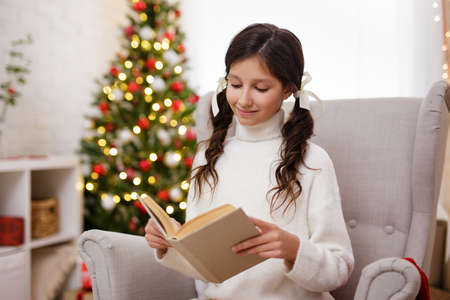 portrait of cute girl reading book in decorated living room with christmas tree