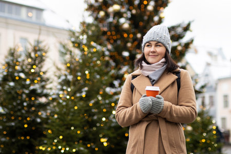 Woman in coat with cup of coffee near christmas trees in old townの写真素材