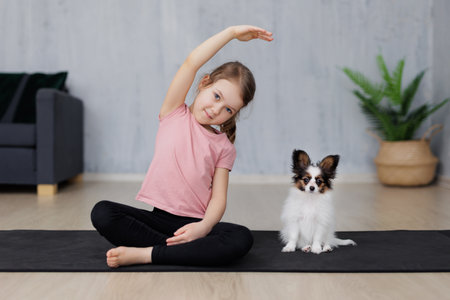cute little girl doing stretching exercises, sitting with her puppy papillon dog on yoga mat at homeの写真素材