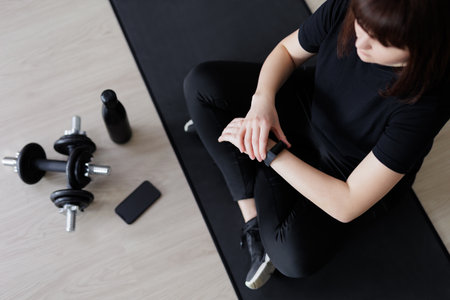 sport and technology concept - top view of young woman sitting on yoga mat, looking at her smart...