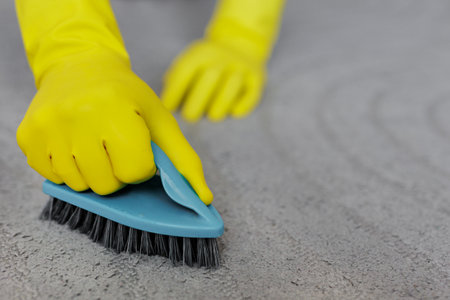 cleaning concept - close up of hands in yellow rubber gloves cleaning grey carpet with brushの写真素材
