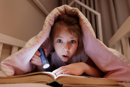 surprised girl lying in bed and reading book with flashlight under blanket in dark roomの写真素材