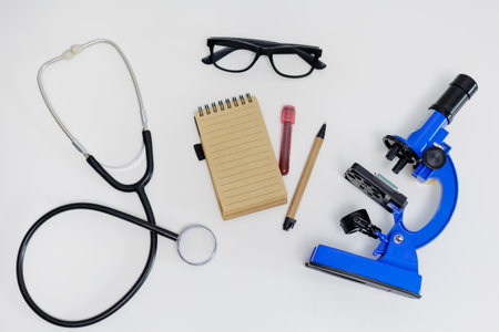 Top view of microscope, stethoscope, eyeglasses and notebook with pen on white