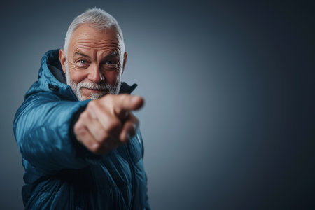 smiling sporty senior man in down jacket pointing forward to camera over bright studio background with copy spaceの素材