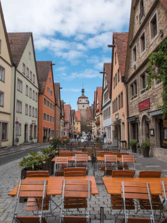 View perspective central street center of medieval town Rothenburg. Side tables and bar chairs in the foreground, side walled houses and tower background with exit door from the fortification wall.のeditorial素材