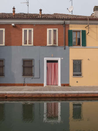 Comacchio, FE, Italy - November 4, 2017: exterior facing room overlooking the canal of Comacchio. Reflection in the water. White entrance door frame with red and white striped curtainのeditorial素材