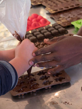 close-up of color hand (adult) that supports a child's hand to make small chocolates with the use of the pastry bag. Rubber molds with various shapes of chocolates.の写真素材