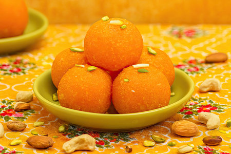 A vibrant and culturally rich image showcasing five orange motichoor ladoos arranged in a pyramid on a green plate, garnished with nuts and surrounded by scattered dry fruits. Set against a festive yellow-patterned cloth, the composition evokes traditional Indian celebrations and religious offerings. Ideal for themes related to Indian cuisine, festivals, cultural food photography, and editorial stock imagery.の素材