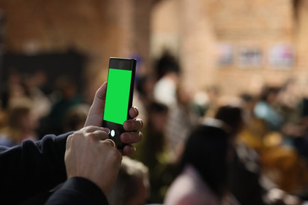 A person is holding a smartphone that features a green screen, surrounded by a vibrant crowd sceneの写真素材