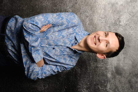 A young man with a bright smile in a stylish blue patterned shirt stands against a dark backgroundの写真素材