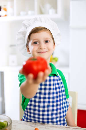 Little cute child boy with cook hat holding tomato in kitchenの写真素材