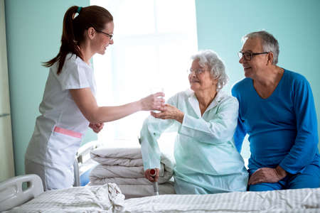 Nursing home, young careful nurse brings water to old woman at nursing homeの写真素材