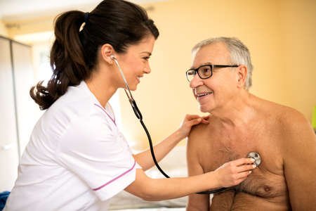Smiling professional feamle doctor examining senior man with stethoscope at nursing homeの写真素材