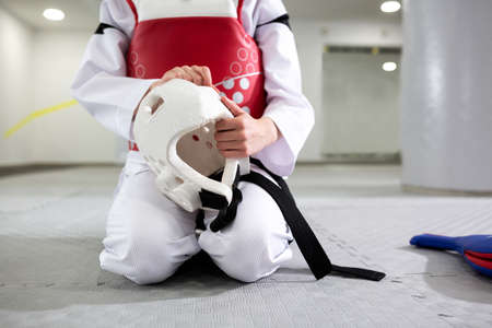 Martial artist in protective gear sitting down and holding a helmet, black belt, white uniform, taekwondoの写真素材