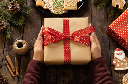 Woman holding a beautifully decorated Christmas gift on rustic wooden table next to a plate full of...