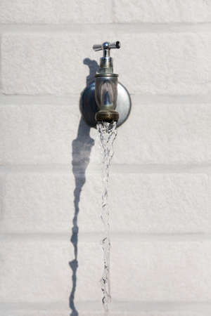 water running from grey metal faucet on a white stone wallの写真素材