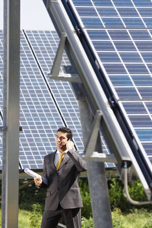 Portrait of mid adult italian male engineer holding blueprints in solar power station and talking on mobile phone. Vertical shape, side view. Copy spaceの写真素材