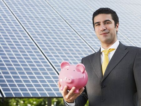 Portrait of mid adult italian male engineer holding piggy bank in solar power station, smiling at camera.Horizontal shape, front view. Copy spaceの写真素材