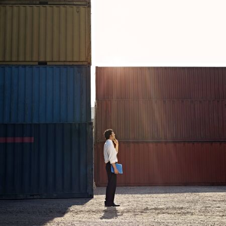Mid adult businessman standing near cargo container with hand on chin and looking up. Square shape,...