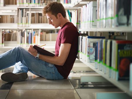 male college student sitting on floor in library, reading book. Horizontal shape, side view, three quarter length, copy spaceの写真素材