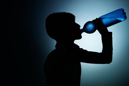 silhouette of young woman drinking water from bottle on blue background. Horizontal shape, side view, copy spaceの写真素材