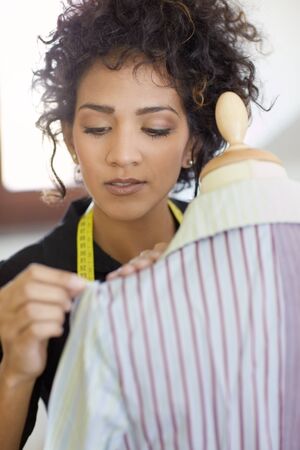 Young hispanic female dressmaker adjusting clothes on tailoring mannequin and smiling. Vertical shape, head and shouldersの写真素材