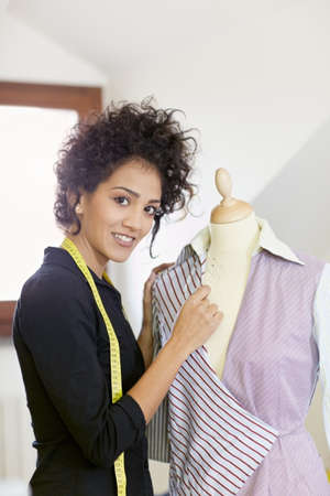 Young hispanic female dressmaker adjusting clothes on tailoring mannequin and smiling at camera. Vertical shape, side view, waist upの写真素材