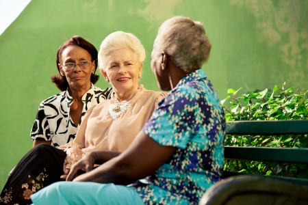 Active retired elderly women and free time, group of happy senior african american and caucasian female friends talking and sitting on bench in parkの写真素材