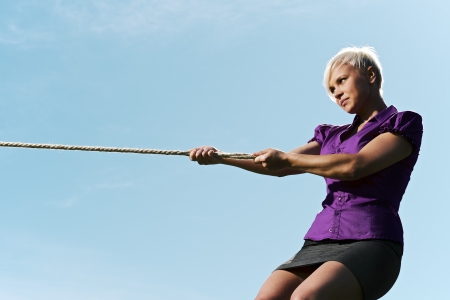 resolute business woman pulling rope against blue skyの写真素材