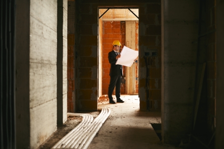Engineer at work in construction site, standing in apartment building and looking at blueprints and plansの写真素材